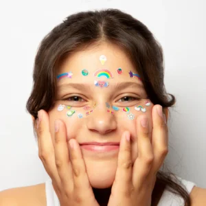 A young person smiles, holding their face with both hands, adorned with Kawaii - Face Stickers in rainbows, stars, hearts, and playful shapes. Perfect inspiration for unique ideas at your favourite gift shop. Plain white background. Gate21.ee