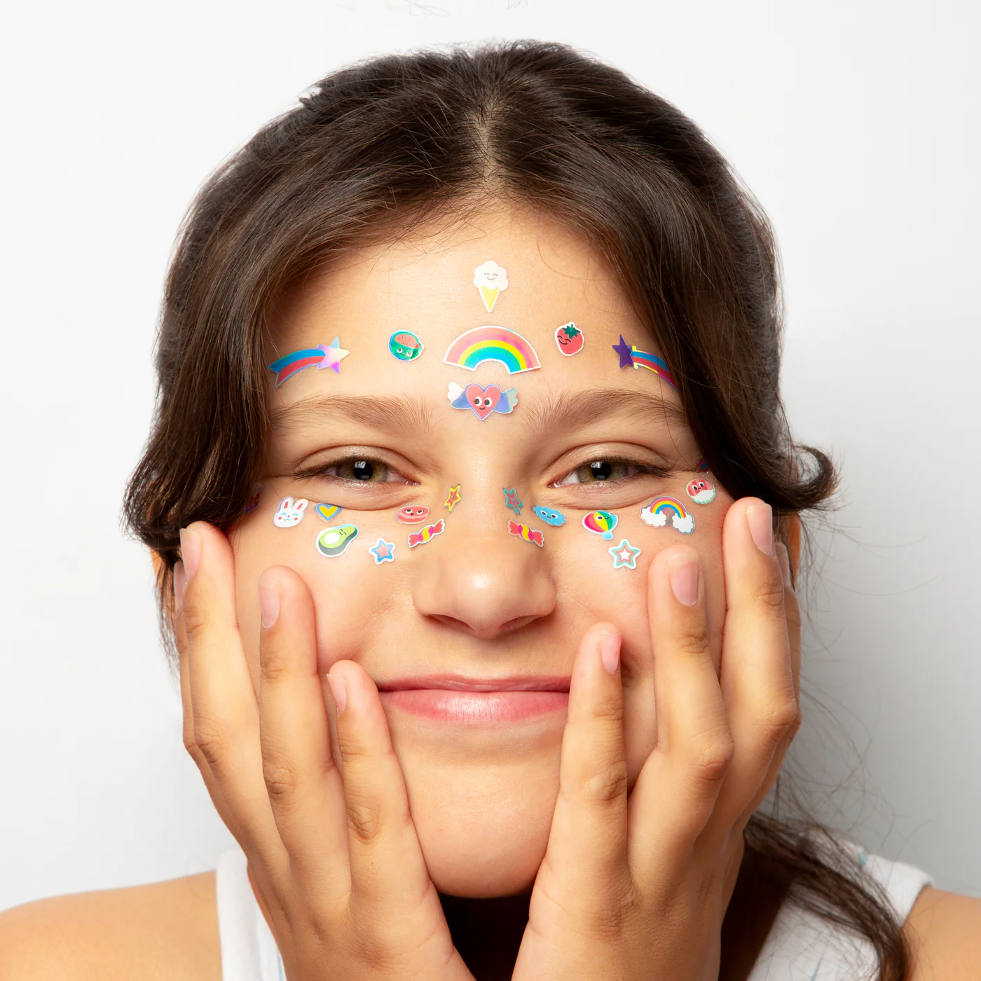 A young person smiles, holding their face with both hands, adorned with Kawaii - Face Stickers in rainbows, stars, hearts, and playful shapes. Perfect inspiration for unique ideas at your favourite gift shop. Plain white background. Gate21.ee