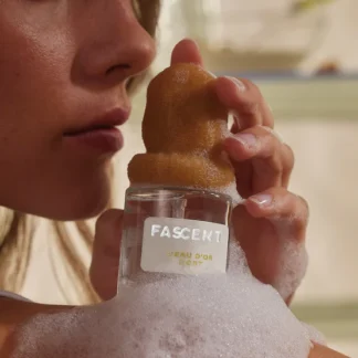 A person holds a 30ml bottle of FASCENT L'eau d'or Port, covered in bubbles with a rounded cap inspired by Estonian design. Their shoulder and part of their face are visible, suggesting the unique concept behind this shop find. Gate21.ee