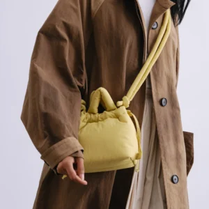 A person wears a brown oversized coat over a white top and cream skirt, carrying the Micro Ona Soft Bag in lime with a knotted strap—an accessory reminiscent of something from a curated gift shop. The photo is cropped just below the chin. Gate21.ee