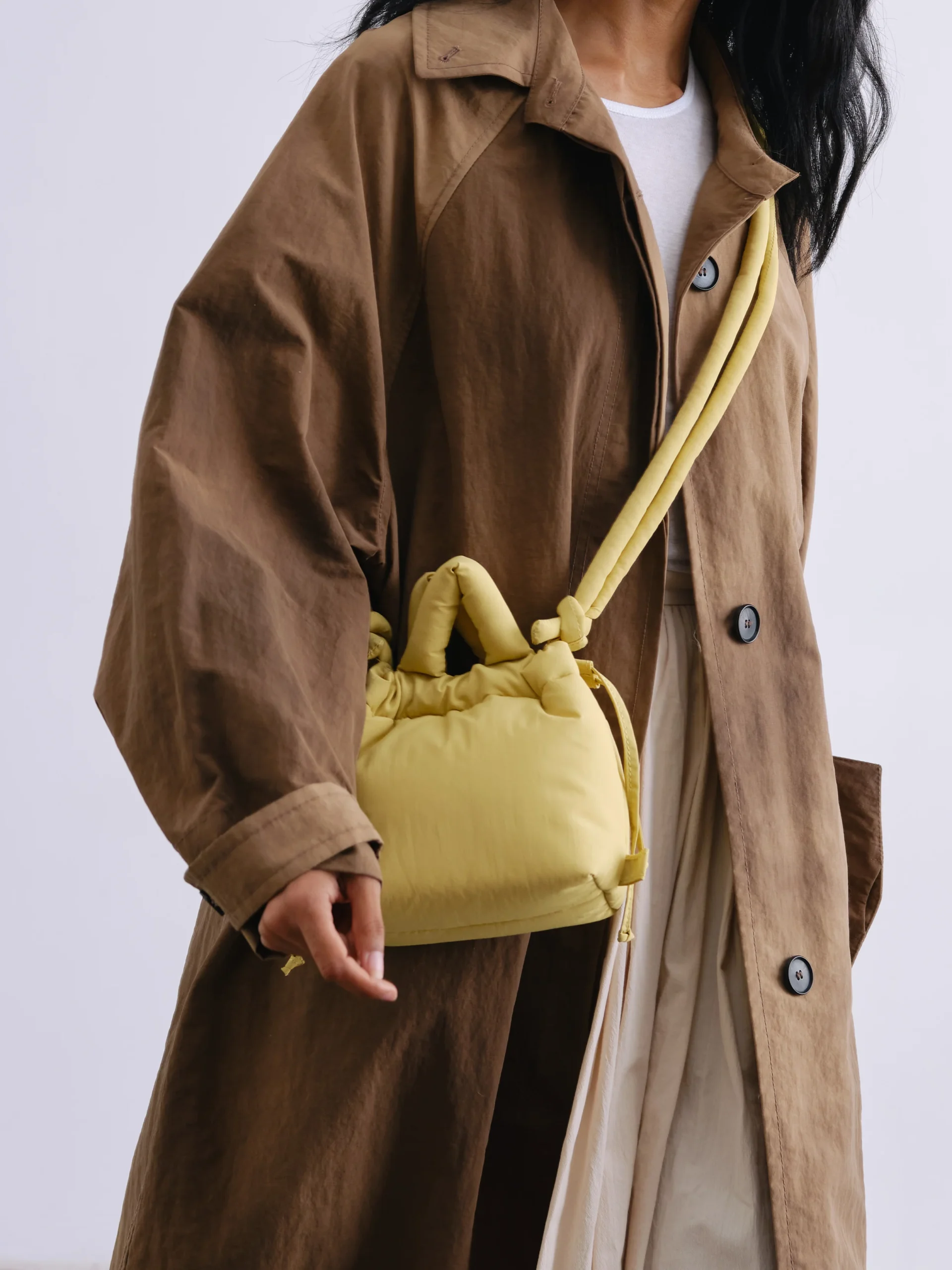A person wears a brown oversized coat over a white top and cream skirt, carrying the Micro Ona Soft Bag in lime with a knotted strap—an accessory reminiscent of something from a curated gift shop. The photo is cropped just below the chin. Gate21.ee