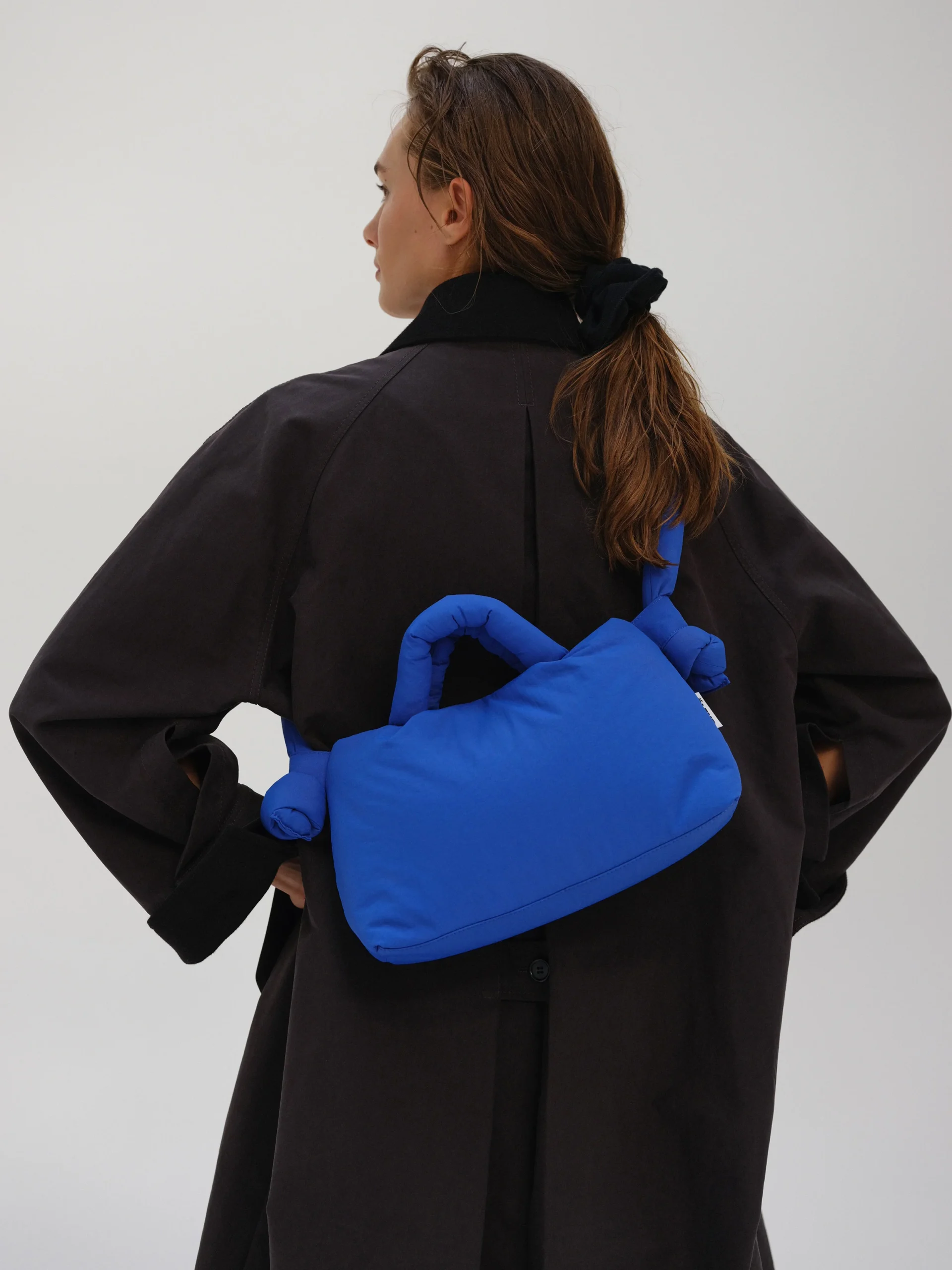 A person with long brown hair tied back, wearing a dark oversized coat, stands with their back to the camera in front of a Tallinn concept shop, carrying the Mini Ona Soft Bag in Cobalt Blue over one shoulder. Gate21.ee