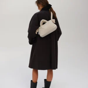 A person stands facing away from the camera in a black coat, black boots, and the Mini Ona Soft Bag - Sand against a plain light background, showcasing modern Estonian style ideal for your next Tallinn shopping trip. Gate21.ee