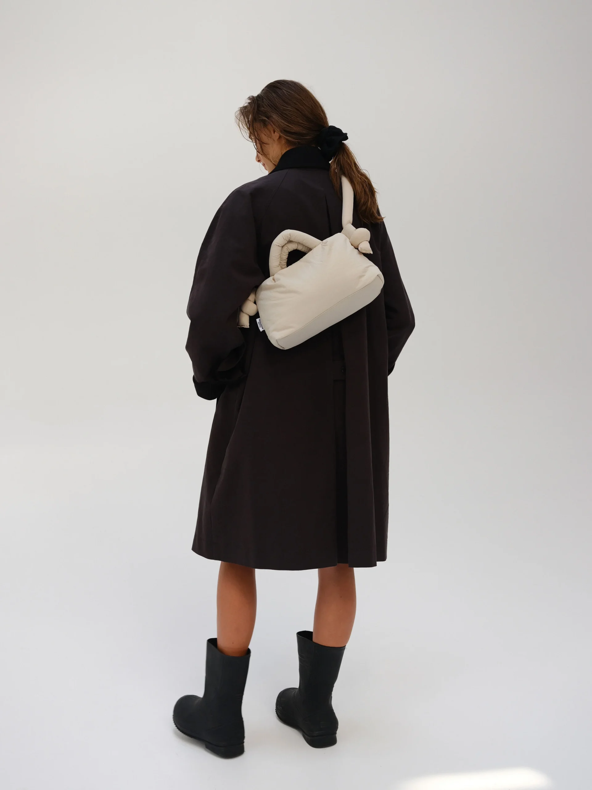 A person stands facing away from the camera in a black coat, black boots, and the Mini Ona Soft Bag - Sand against a plain light background, showcasing modern Estonian style ideal for your next Tallinn shopping trip. Gate21.ee