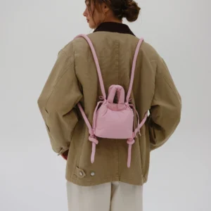 A person in a tan oversized jacket and light trousers is seen from behind, carrying the Micro Ona Soft Bag in Pale Pink. The plain white background evokes the clean aesthetic of a modern concept shop. Gate21.ee