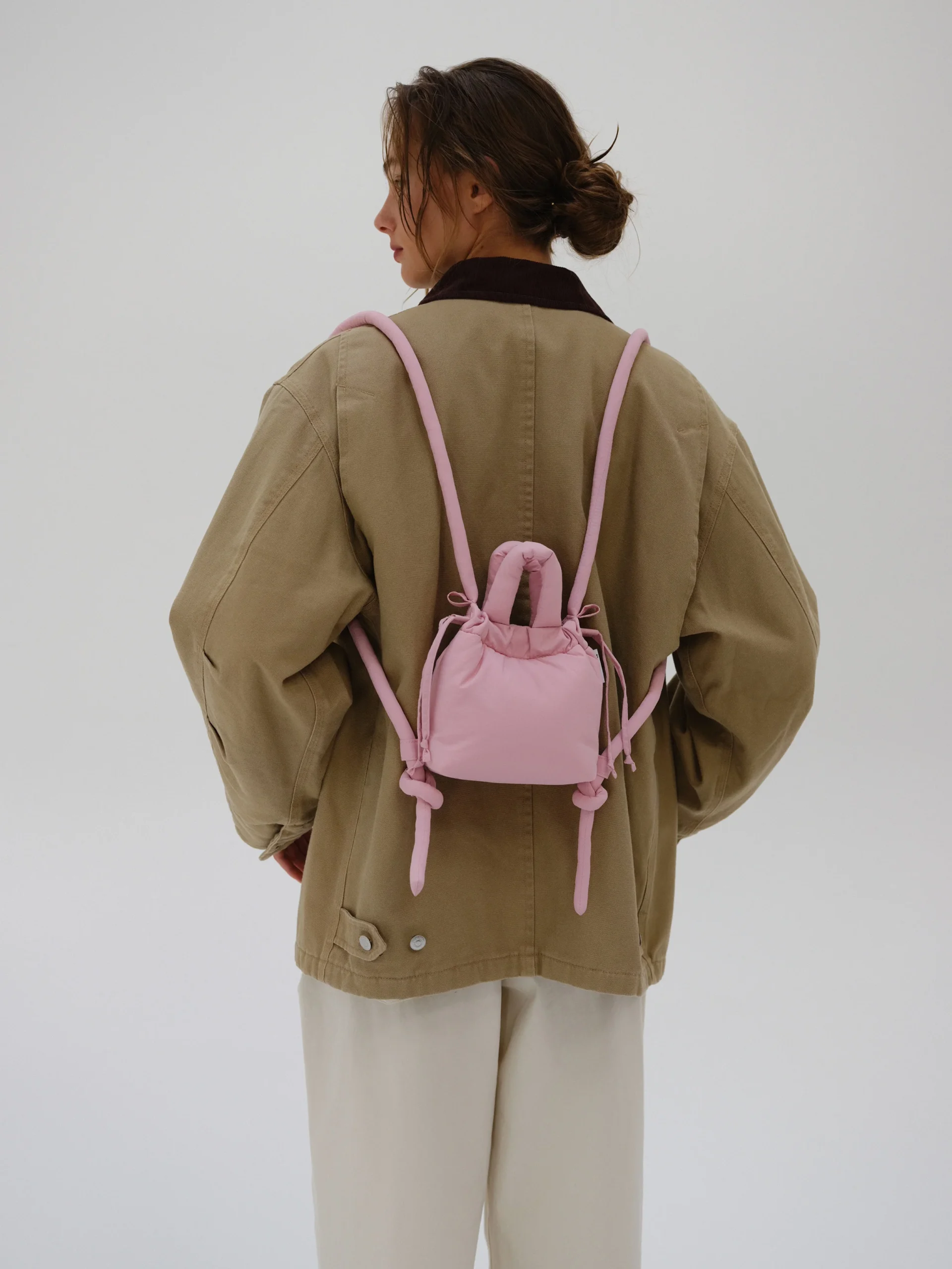 A person in a tan oversized jacket and light trousers is seen from behind, carrying the Micro Ona Soft Bag in Pale Pink. The plain white background evokes the clean aesthetic of a modern concept shop. Gate21.ee