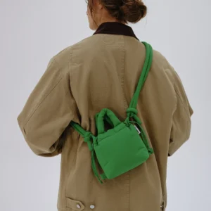A person with brown hair tied back wears a tan jacket, white trousers, and carries the Micro Ona Soft Bag in green—ideal for trips to curated concept shops or chic gift shops. The background is plain and light-coloured. Gate21.ee
