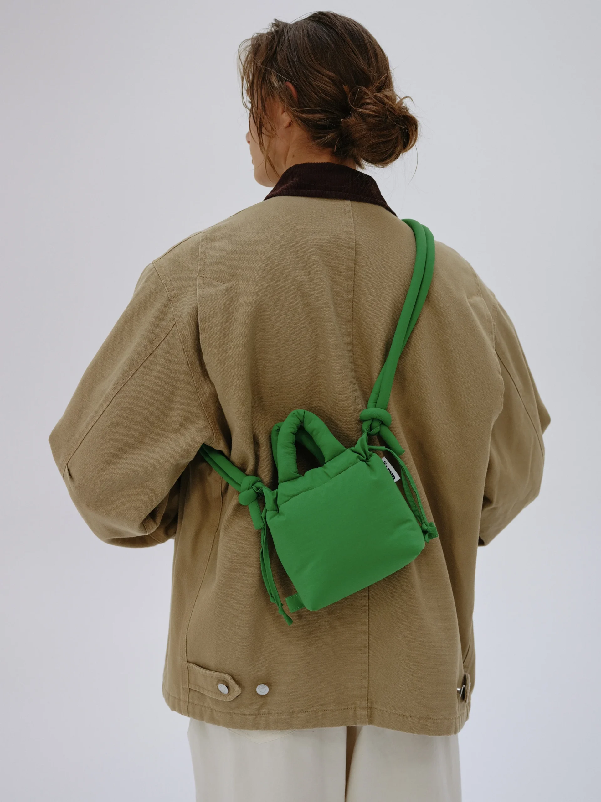 A person with brown hair tied back wears a tan jacket, white trousers, and carries the Micro Ona Soft Bag in green—ideal for trips to curated concept shops or chic gift shops. The background is plain and light-coloured. Gate21.ee