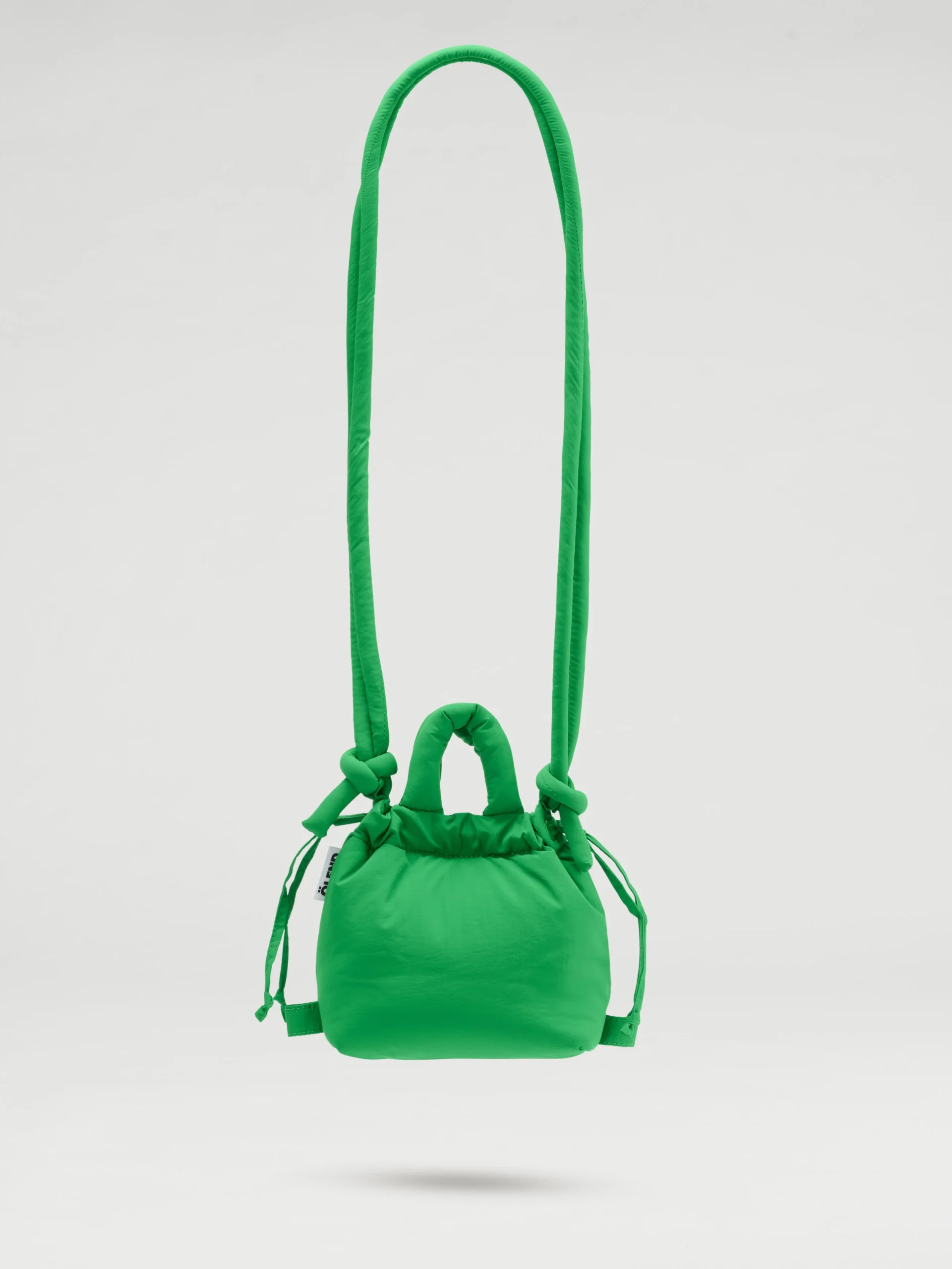 The Micro Ona Soft Bag - Green is a compact, vibrant handbag featuring a long padded strap and knotted accents. This curated piece of Estonian design is displayed suspended against a plain white background. Gate21.ee