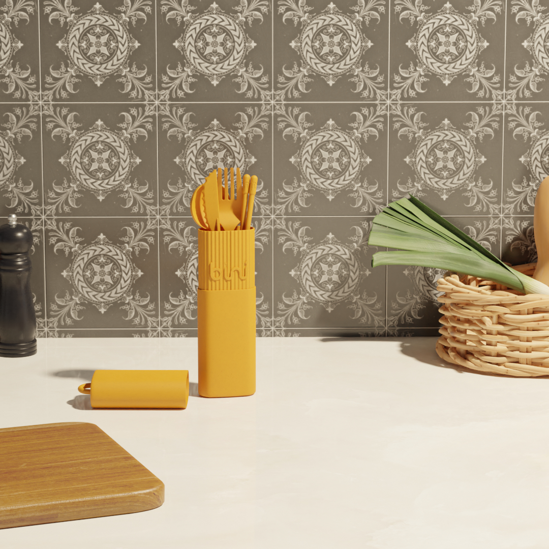 The Bini Cutlery Kit - Yellow sits in its matching case on a light worktop, with patterned grey tiles, a chopping board, a leek, a wicker basket, and a salt cellar nearby. Gate21.ee