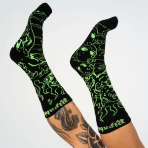 Wearing Horntail Athletic Socks - Black, a person with a leg tattoo showcases black socks featuring bright green Estonian-inspired designs of creatures and abstract patterns, all set against a white background. Gate21.ee