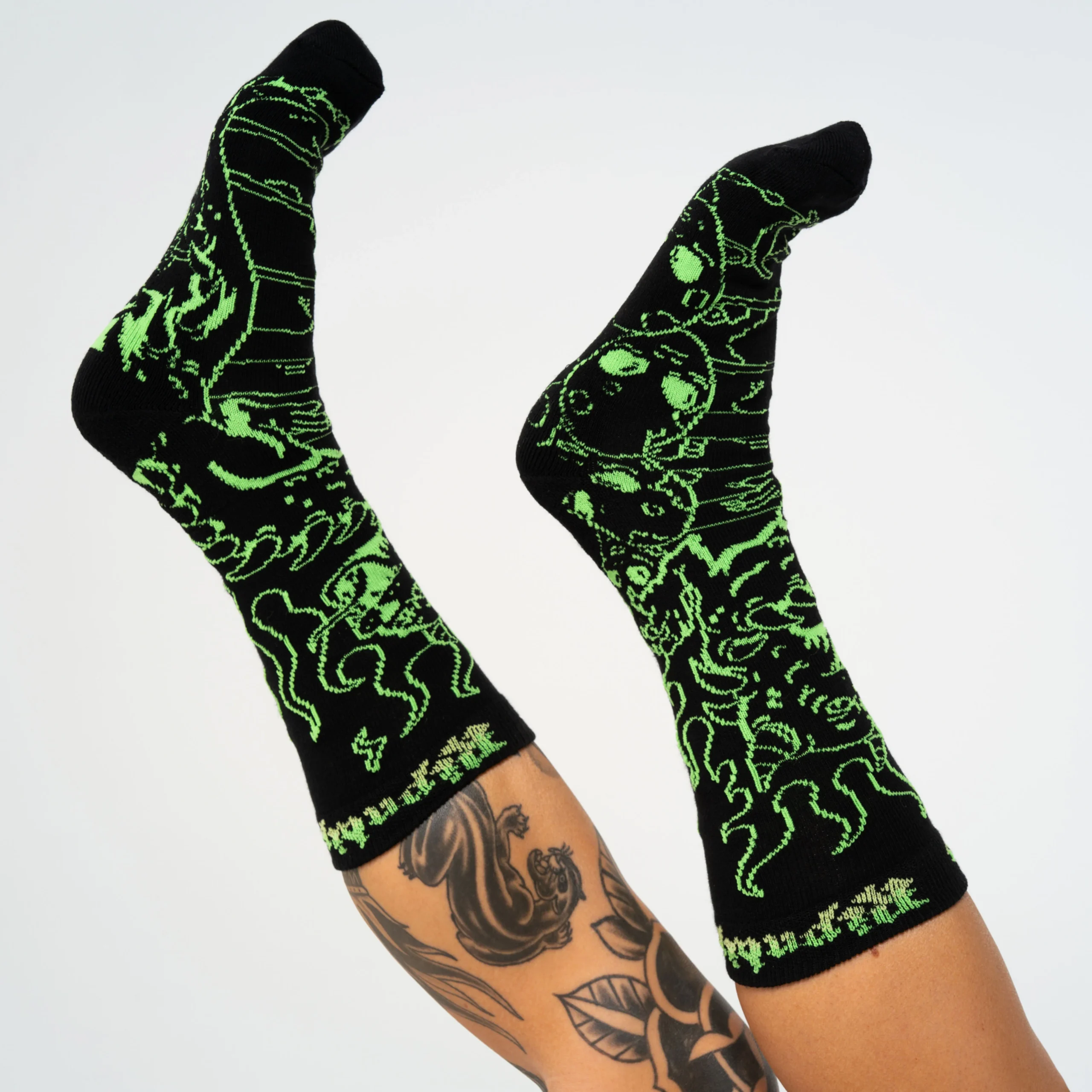 Wearing Horntail Athletic Socks - Black, a person with a leg tattoo showcases black socks featuring bright green Estonian-inspired designs of creatures and abstract patterns, all set against a white background. Gate21.ee