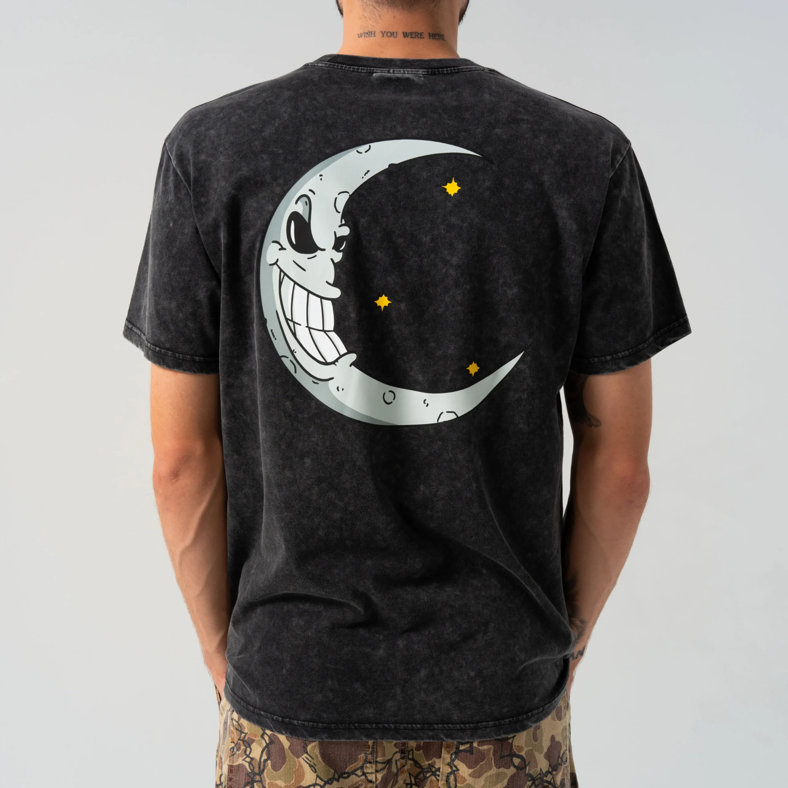 A person wears the Oracle Tee - Black Wash, featuring a large cartoon crescent moon and yellow stars on the back—ideal for Tallinn shopping or as a unique gift shop find. The smiling moon faces away as the wearer shows their back to the camera. Gate21.ee