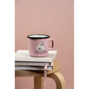 The Together - Moomin pink enamel mug 3.7dl, featuring a cartoon and floral design, sits on a closed beige notebook atop stacked books on a wooden stool, all set against a plain pink background. Gate21.ee