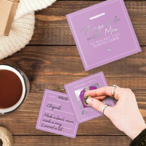 A hand scratches a purple Mum - Scratch Card beside tea, a notepad, and a "Things to do with Mum" booklet—ideal for discovering Estonian design treasures or unique gifts when shopping in Tallinn’s local gift shops. Gate21.ee