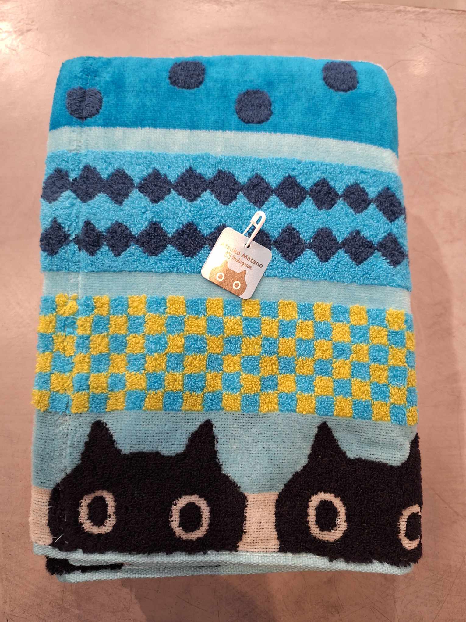 The Bath Towel - KÕIK - 120cm features blue, turquoise, and yellow geometric patterns with dots, diamonds, and chequerboards, plus a row of black cat faces at the bottom—a playful Estonian design with a tag on one corner. Perfect for gift shops. Gate21.ee