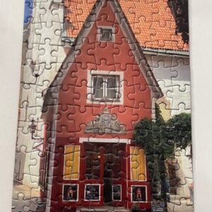 Discover the Red House - Micro Puzzle from our gift shop: a finished jigsaw featuring a small, red house with a triangular tiled roof, yellow window shutters, lush plants, and a pale background. Gate21.ee