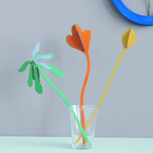 Bouquet Autumn Tango features three vibrant paper cut-out plants—green, orange, and yellow—arranged in a clear glass against a light grey wall with a blue circular frame, ideal for unique displays in concept shops or gift shops. Gate21.ee
