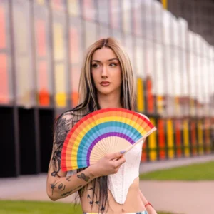 A person with long, straight hair and visible tattoos holds the "Rainbow" fan, a vibrant piece of Estonian design, whilst standing outdoors in front of a modern glass building reflecting colourful light. Gate21.ee