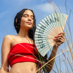 Wearing a red bandeau top, a woman with long plaits and gold hair accessories holds the "Super fan!" fan outdoors, eyes closed as if gathering creative gift shop ideas, with grass in the foreground. Gate21.ee