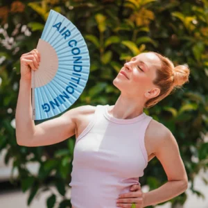 A woman outdoors smiles with eyes closed while fanning herself with the "Air Conditioning!" fan amid green leaves—inspired by curated Estonian design. Gate21.ee