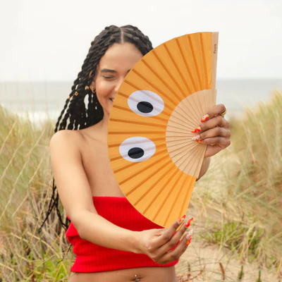 A woman stands outdoors in tall grass, holding the playful "Eyes" fan—an orange accessory with cartoon eyes—capturing the spirited essence of Estonian design and offering fresh inspiration for Tallinn shopping. Gate21.ee