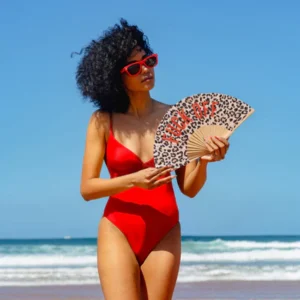 Wearing a red swimming costume and red sunglasses, a person stands on the beach holding the "Fuck off!" leopard fan—a bold accessory for those who love standout gift shop finds. Behind them, clear skies meet shimmering ocean waves. Gate21.ee