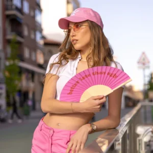 A woman stands outdoors holding the "Better late than ugly!" fan, wearing a pink cap, pink sunglasses, white cropped top, and pink shorts—an eye-catching item ideal for any gift shop or concept store. Gate21.ee