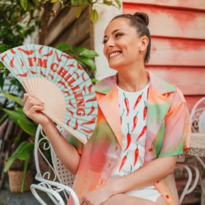 Outdoors, a woman smiles from a white chair, holding the “I’m chilling” fan—featuring red chilli peppers—from a curated concept shop. She pairs a colourful shirt with a white dress adorned in chilli pepper prints. Gate21.ee