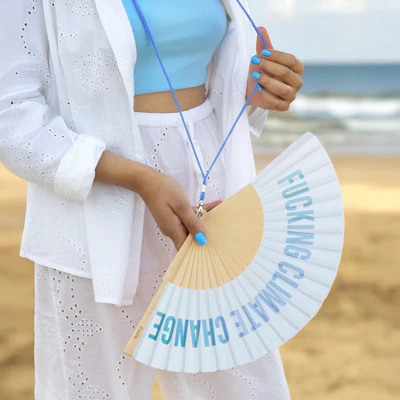 Wearing a white broderie anglaise outfit, someone holds the "Climate Change!" fan with bold blue text, their blue nails adding flair—ideal for finding unique picks on your next Tallinn shopping spree or at a gift shop. Gate21.ee