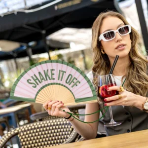 A woman in white sunglasses holds a cocktail and the "Shake it off!" fan whilst relaxing at an outdoor café table, enjoying Tallinn shopping after discovering unique finds at a local concept shop. Gate21.ee