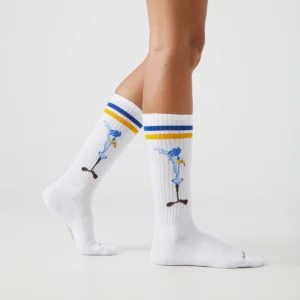 A person models the Duffy & Bugs Bunny – Athletic white knee-high socks with blue and yellow stripes and a cartoon Road Runner character, standing on a plain white surface. Gate21.ee
