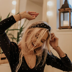 A woman with long blonde hair, a black hairband, and a spotted black top smiles joyfully with her eyes closed and arms raised—radiating the happiness of finding Queen. A mirror with lights and a lantern are visible behind her. Gate21.ee