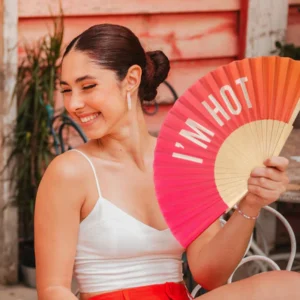 A woman with dark hair in a bun, wearing a white top and red shorts, smiles outdoors whilst holding the "I'm hot!" - gradient fan from a concept shop; the fan features bold white text on a red and orange background. Gate21.ee