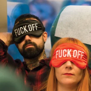 A tattooed, bearded man and a red-haired woman wear boldly printed "Fuck off!" - sleeping masks&mdash;an edgy Estonian design&mdash;while sitting side by side, possibly on public transport. Gate21.ee