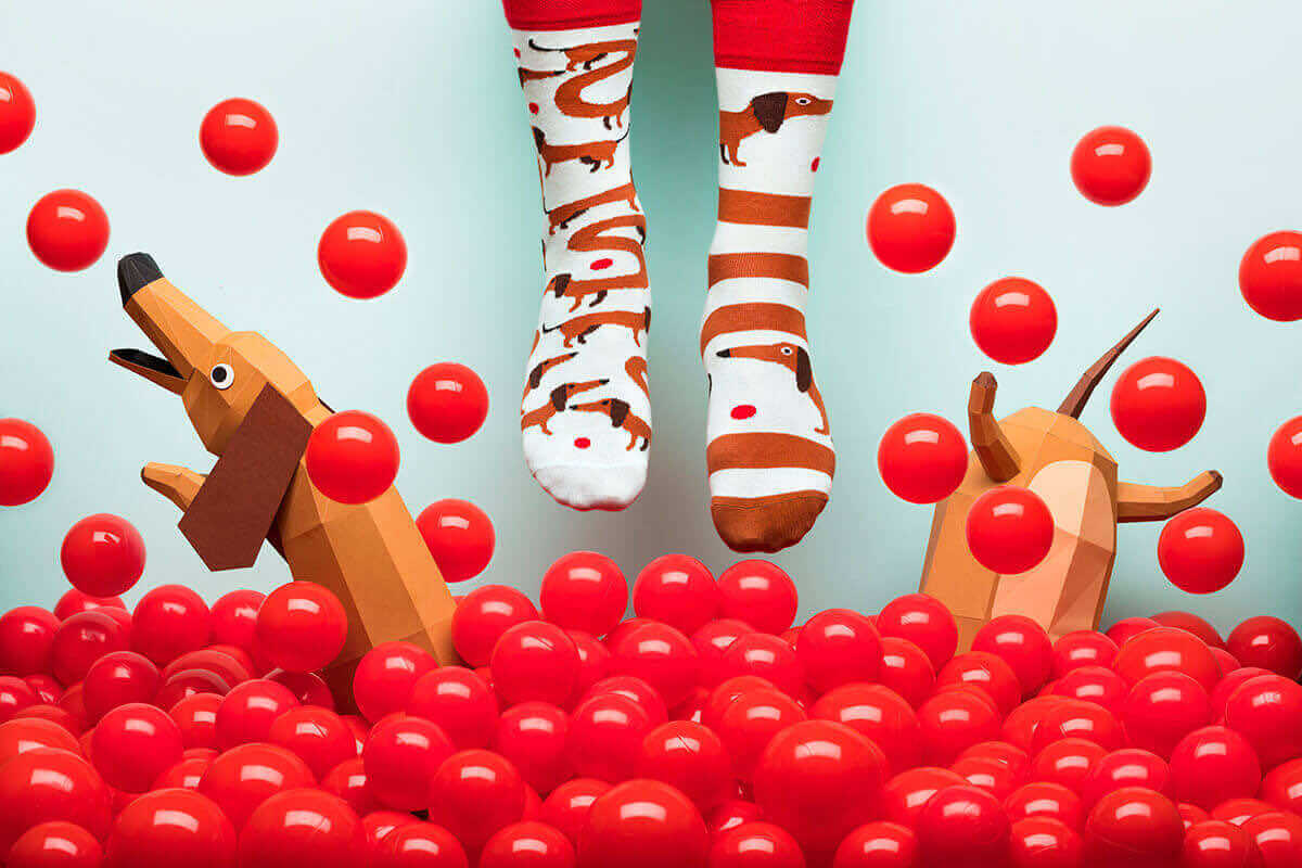 A pair of feet in colourful Sausage Dog socks with dachshund designs dangle above red plastic balls. Two geometric dog sculptures, inspired by Estonian design, add a playful touch—perfect for unique gift shop ideas. Gate21.ee