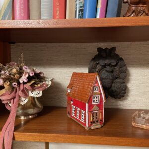 A wooden shelf displays the Red House - Tea light, a decorative vase with dried flowers and pink ribbon, a dark owl-shaped wall ornament, and a clear glass box&mdash;unique gift ideas for any shop. Books can be seen on the shelf above. Gate21.ee