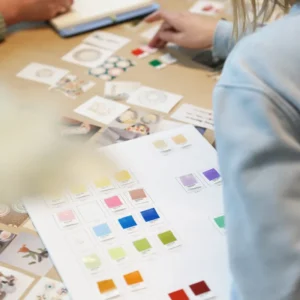 Several people sit at a table covered with curated colour swatches, printed images, and notes as they discuss the Bottle Stopper Tulip - Yellow (7 x 3.5 x 12 cm), reviewing its samples for upcoming Tallinn shopping trends. Gate21.ee