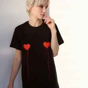 A person with short blonde hair wears the LUKS - 2 S&uuml;da - T-shirt, a stylish black T-shirt featuring two red heart designs with strings on the chest, standing against a light background. Gate21.ee