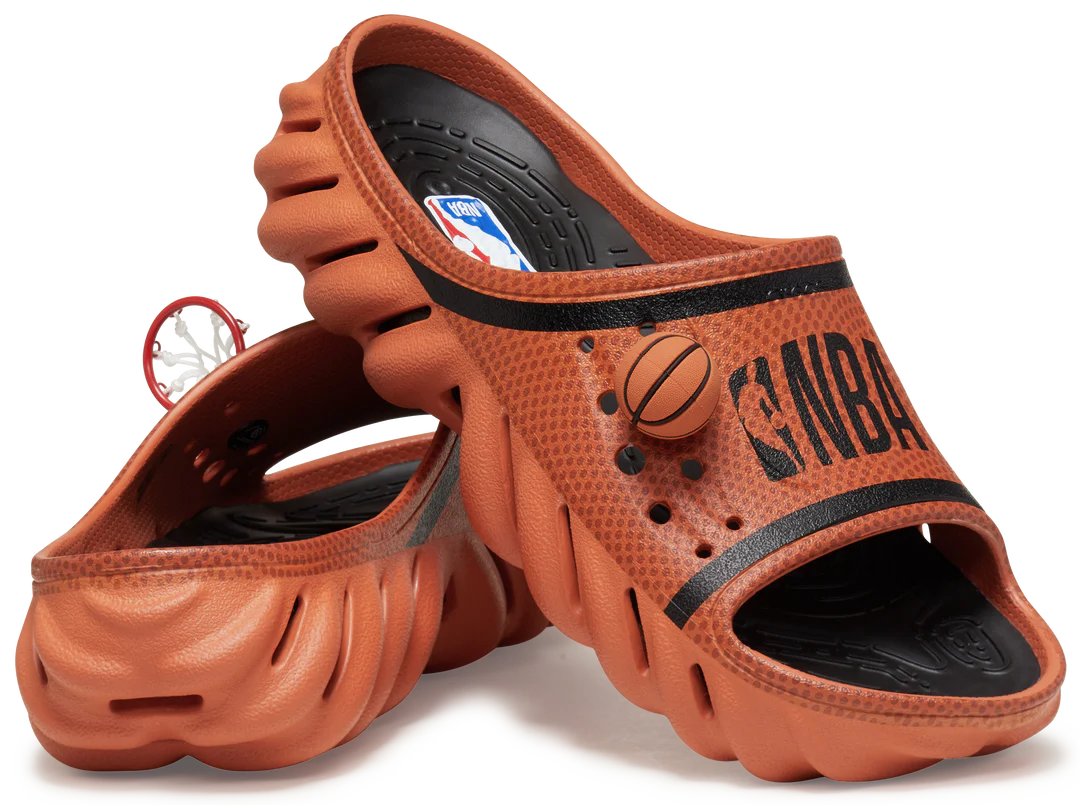 The Crocs™ NBA Echo Slide in Sienna features an orange, basketball-textured slip-on design with NBA branding and wavy soles—ideal for gift shops or concept stores. One slide stands upright whilst the other rests on its side. Gate21.ee