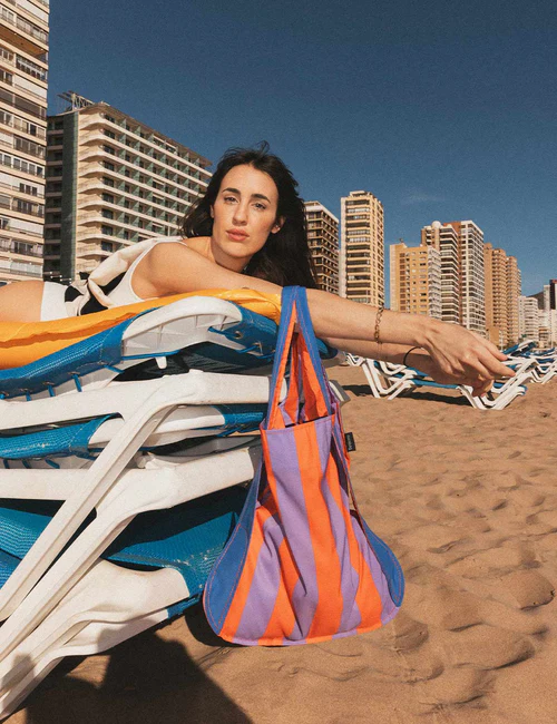 A woman with dark hair in a patterned swimming costume lounges on stacked sunbeds in Tallinn, her Royal Stripes - Special Edition bag with orange and purple Estonian design hanging nearby under the clear blue sky. Gate21.ee