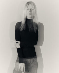 A woman with long blonde hair wears the LUKS - 2 S&uuml;da - T-shirt, jeans, and a dark long-sleeve top against a light background. The double exposure effect creates overlapping, semi-transparent outlines of her. Gate21.ee