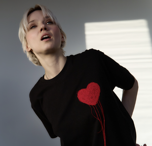 A person with short blond hair wears the LUKS - 1 Süda - T-shirt, a black tee featuring a bold red heart. Standing by a sunlit wall, they look upward, effortlessly highlighting this trendy shirt’s unique style. Gate21.ee