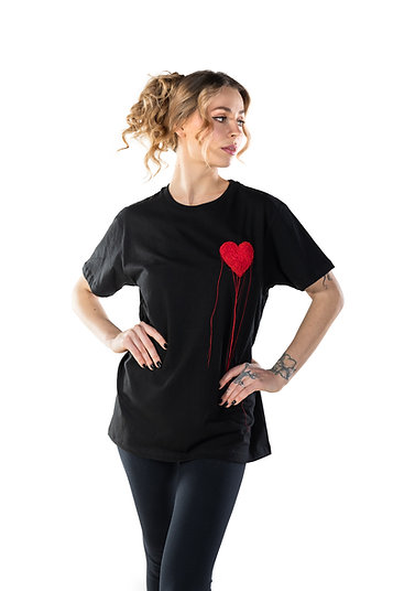 A woman with light brown curly hair wears the LUKS - 1 Süda T-shirt, a black shirt featuring a red dripping heart—a standout Estonian design. She poses confidently against a white background—ideal for unique Tallinn shopping finds. Gate21.ee