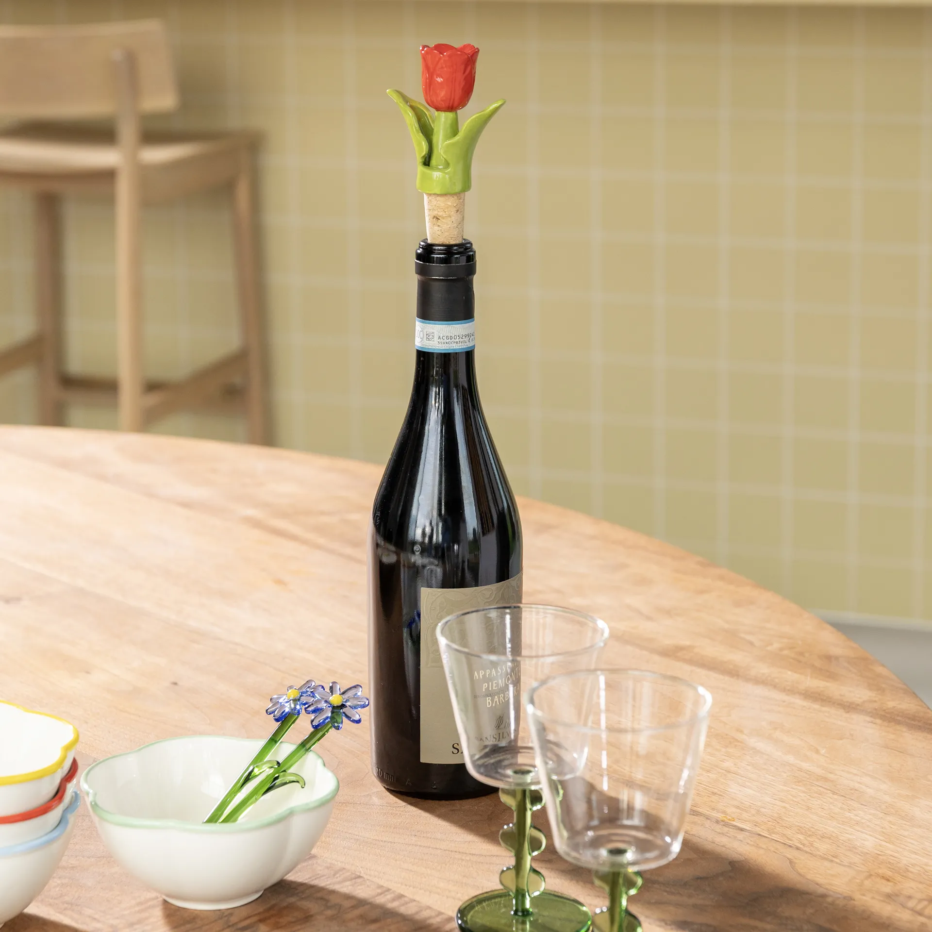 A Bottle Stopper Tulip - Red (7 x 3.5 x 12 cm) decorates a wine bottle on a wooden table, accompanied by two empty glasses and stacked bowls—ideal for anyone seeking unique Tallinn shopping or gift shop finds. Gate21.ee