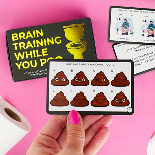 A hand holds a card featuring eight cartoon poo emojis labelled A to H, each with a unique expression. In the background is "Brain Training whilst you Poo - Cards," a quirky Estonian design game spotted in a Tallinn concept store. Gate21.ee