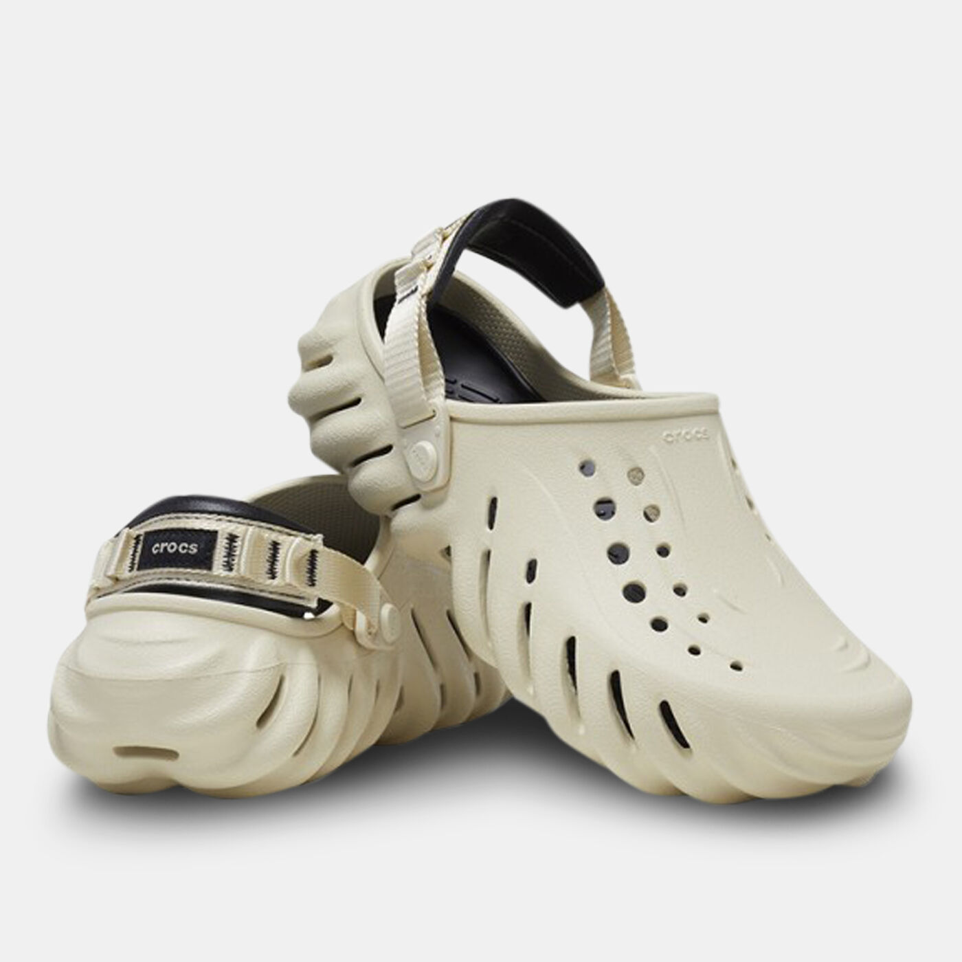 A pair of Crocs™ Echo Clog in Bone/Black, featuring black adjustable heel straps, ventilation holes on top, and a wavy textured sole. One shoe stands upright whilst the other is tilted on its side. Estonian design inspired. Gate21.ee