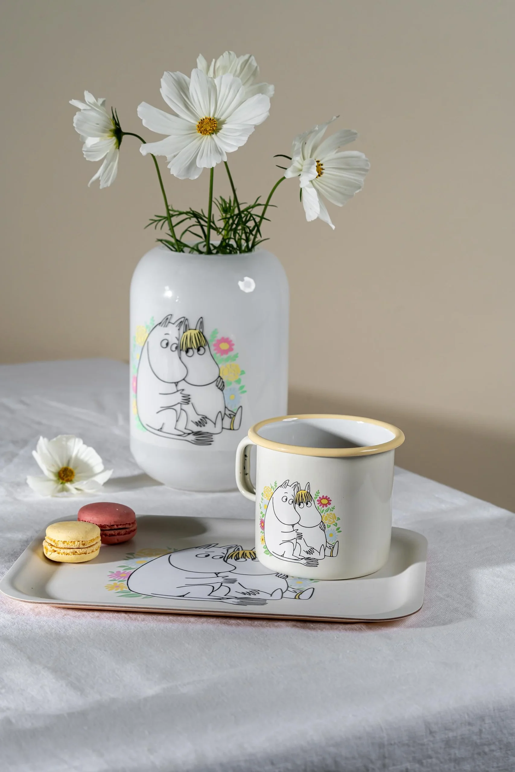The Flowers - Moomin enamel mug 550ml, adorned with charming Moomin designs, sits on a white tablecloth beside macarons and a single white flower—ideal for fans of Estonian design. Gate21.ee
