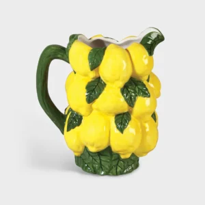 The Jug Lemon (18 x 13 x 20.5 cm) is a ceramic jug shaped and painted like bright yellow lemons with green leaves and handle, adding a playful splash of colour to any gift shop or concept store. Shown on a plain light grey background. Gate21.ee
