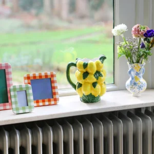 A curated windowsill display features three empty gingham photo frames, a Jug Lemon 18 x 13 x 20.5 cm, and a floral vase with assorted blooms, evoking the charm of an Estonian design shop against a blurred outdoor view. Gate21.ee